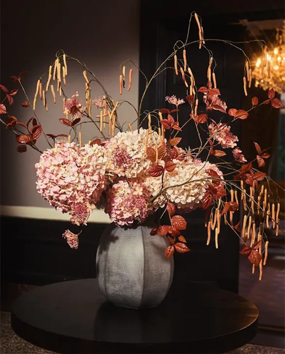 Here is a stunning bouquet of silk flowers from Silk-ka's autumn collection, featuring an elegant and refined composition. The pink hydrangeas, combined with branches adorned with autumnal leaves and delicate catkins, create a warm and sophisticated atmosphere. This bouquet, placed in a sleek vase, perfectly exemplifies the exceptional quality and realism of Silk-ka's creations.