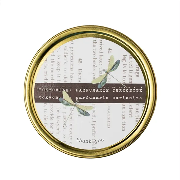 Exquisite Tokyo Milk candle tin featuring a refined vintage-inspired design. The lid displays delicate dragonfly illustrations against a backdrop of antique text. A brown band across the center reads 'TOKYOMILK PARFUMARIE CURIOSITE'. The words 'thank you' are elegantly inscribed at the bottom, all encased in a luxurious gold rim, embodying the brand's signature blend of whimsy and sophistication.