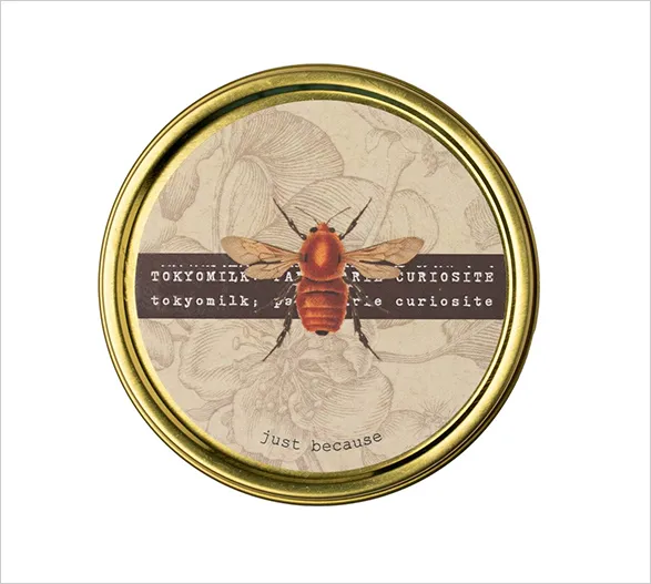 Elegant Tokyo Milk scented candle tin featuring a vintage-inspired design with a vibrant red bee illustration. The label reads 'Tokyo Milk Curiosite' and 'Just because', evoking sophistication and intrigue. The luxurious gold rim adds a touch of opulence to this artisanal beauty product.