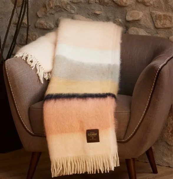 Elegant Mantas Ezcaray throw blanket draped over a brown leather armchair. This Spanish-made luxurious throw features a soft color palette of cream, beige, gray, and peach in gentle stripes. Adorned with delicate fringe, it showcases the brand's renowned craftsmanship. The Ezcaray label is visible, affirming its authenticity. This piece exemplifies Mantas Ezcaray's century-old tradition of creating exquisite, high-quality textiles that blend timeless elegance with modern design.