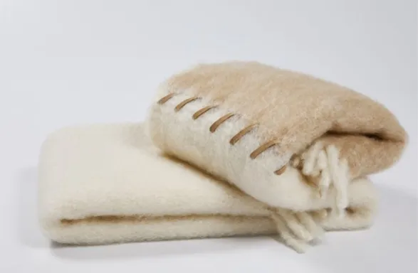 Luxurious throw blanket from Mantas Ezcaray's Stitching Collection. Crafted in Spain, this exquisite piece showcases a creamy off-white hue and delicate fringe detailing. The visible stitching along one edge highlights the brand's commitment to artisanal craftsmanship. Made from premium natural fibers, likely mohair or wool, it embodies Mantas Ezcaray's century-old tradition of creating sumptuous, durable textiles with a contemporary aesthetic.