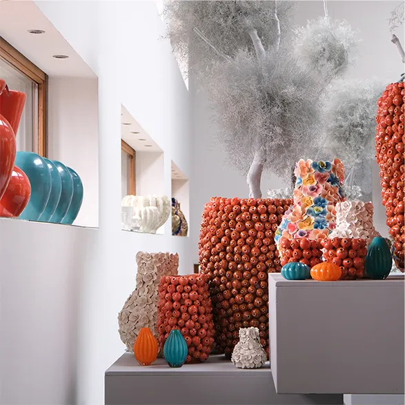 Striking display of Des Pots' handcrafted vases. Vibrant orange vessels adorned with intricate bead-like textures dominate the scene. Smaller teal and multicolored pieces add contrast. Whimsical cloud-like sculptures float above, creating an enchanting atmosphere. This Dutch brand, founded in 1989, showcases its unique style through these eye-catching, versatile art pieces that transform any space into a gallery-like setting.