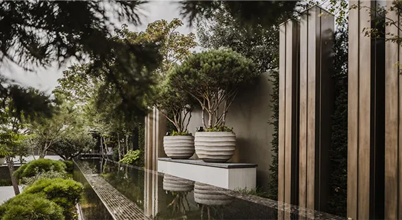 Refined garden vista featuring Atelier Vierkant's signature planters. Sleek, round clay pots in soft grey tones adorn a modernist terrace, reflecting in a serene water feature. The Belgian brand's artisanal craftsmanship seamlessly blends contemporary design with natural elements.
