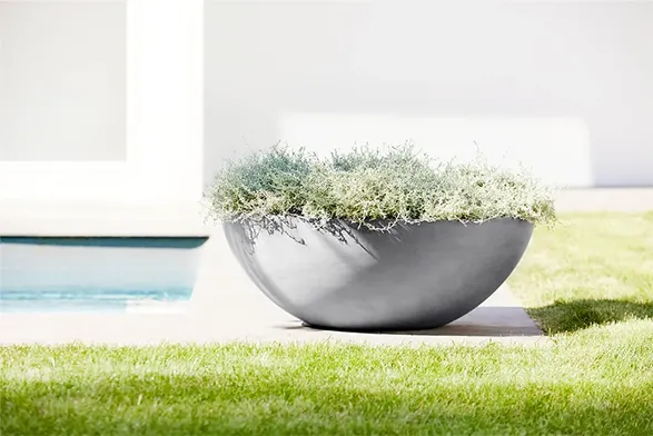 Elegant Ecopots planter: A large, sleek bowl-shaped container in matte gray, filled with lush greenery. Set on a manicured lawn with a pool glimpse. Eco-friendly, made from recycled plastics, combining modern design with sustainability.
