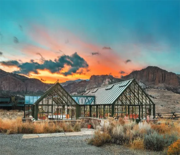 Elegant Hartley Botanic greenhouse gleaming at sunset, nestled in a rugged desert landscape. The glass structure reflects the vibrant orange sky, its classic Victorian design a testament to timeless craftsmanship. Surrounded by arid terrain and distant mesas, this greenhouse embodies Hartley Botanic's commitment to perfect design and enduring strength in even the most challenging environments.