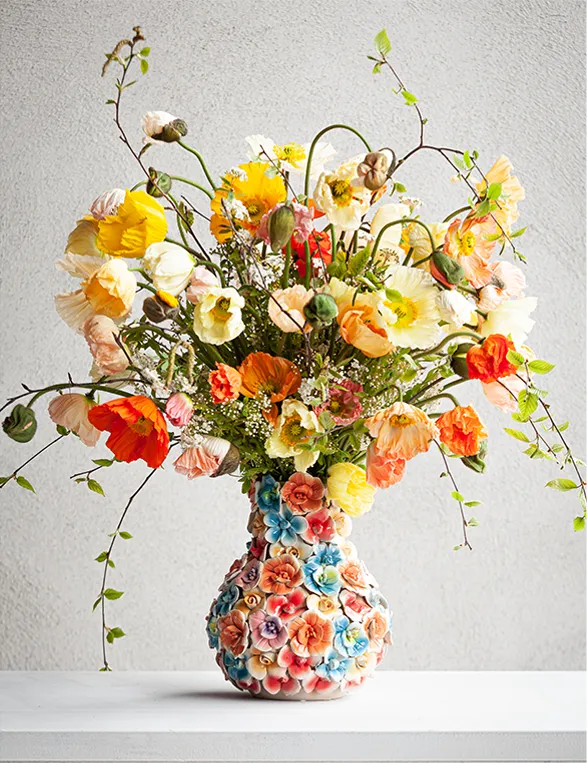 A stunning Des Pots vase showcases its handcrafted uniqueness with a whimsical floral motif in pastel hues. This Dutch brand's commitment to artisanal excellence is evident in the vase's intricate details. Atop, a vibrant Silk-ka arrangement bursts with life-like silk flowers in sunny yellows, fiery oranges, and delicate whites. The interplay between the sculptural vase and the exuberant bouquet creates a captivating centerpiece, embodying the harmony of Dutch design sensibilities.